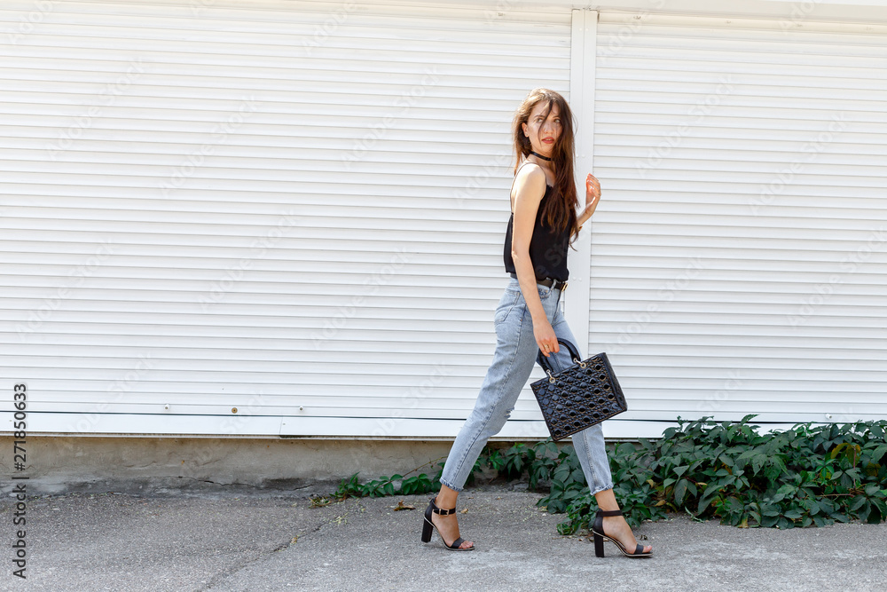 Young stylish woman wearing black cami top, blue cropped denim jeans, black high heel sandals and holding bag walking in the city street. Trendy casual outfit for summer or spring. Street fashion.
