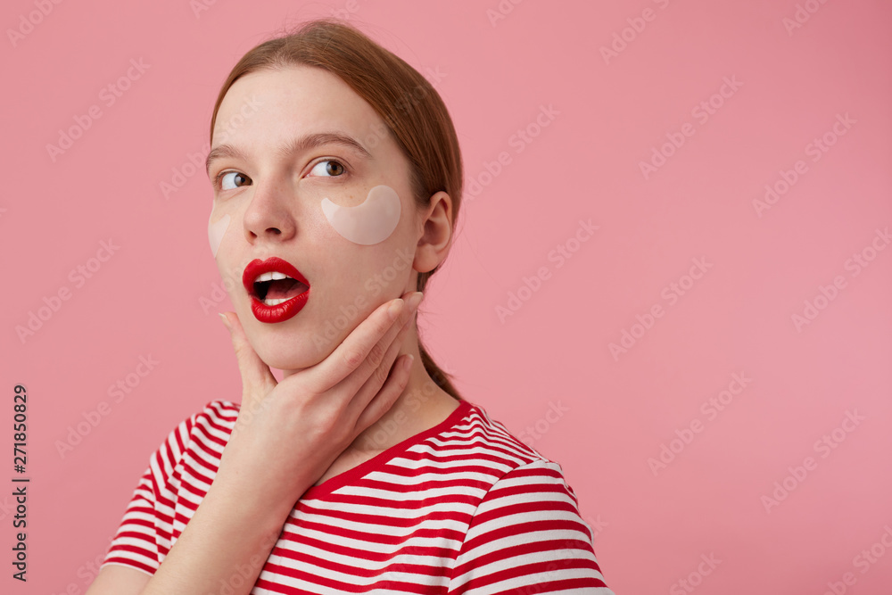 Portrait of cute thinking young red-haired girl with red lips and with patches under the eyes, wears in a red striped T-shirt, looks away, touches the cheek, stands over pink background. Portrait of cute thinking young red-haired girl with red lips and with patches under the eyes, wears in a red striped T-shirt, looks away, touches the cheek, stands over pink background.