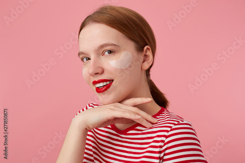 Nice smiling red-haired girl wears in a red striped T-shirt, with red lips and with patches under the eyes, touches the cheen, stands over pink background and enjoying free time for skin care.