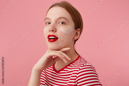 Beauty smiling red-haired lady wears in a red striped T-shirt, with red lips and with patches under the eyes, touches the cheen, stands over pink background and enjoying free time for skin care.