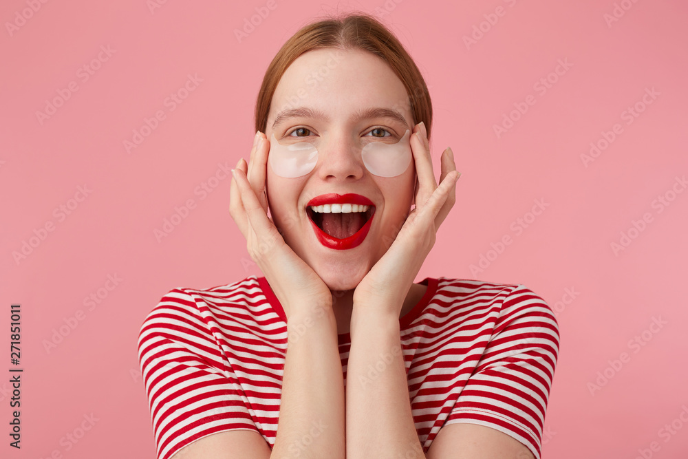 Nice young happy red-haired lady in a red striped T-shirt, with red lips , touches his face with fingers, very pleased with new patches. Stands over pink background. Nice young happy red-haired lady in a red striped T-shirt, with red lips , touches his face with fingers, very pleased with new patches. Stands over pink background.