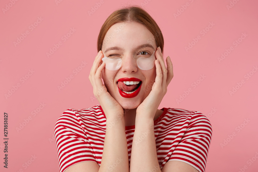 Portrait of cute young winking red-haired lady with patches under the eyes, with red lips and , wears in a red striped T-shirt, looks at the camera and shows tongue , stands over pink background. Portrait of cute young winking red-haired lady with patches under the eyes, with red lips and , wears in a red striped T-shirt, looks at the camera and shows tongue , stands over pink background.