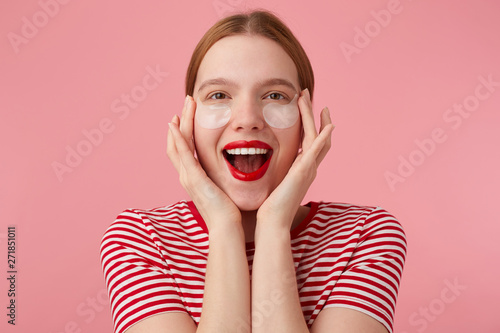 Nice young happy red-haired lady in a red striped T-shirt, with red lips , touches his face with fingers, very pleased with new patches. Stands over pink background.