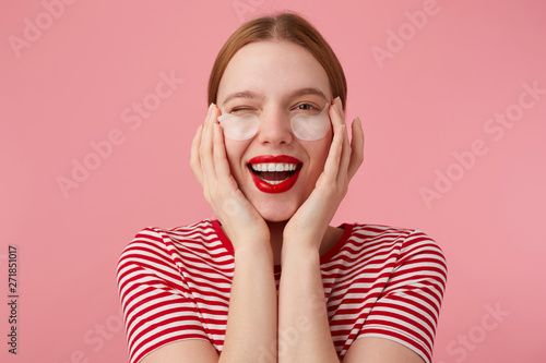 Cute young happy red-haired lady in a red striped T-shirt, with red lips , very pleased with new patches, wink and broadly smiling, touches his face with fingers. Stands over pink background.