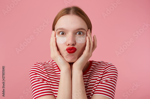 Portrit of funny nice young happy red-haired lady in a red striped T-shirt, with red lips , touches his face with palms, very pleased with new patches. Stands over pink background.