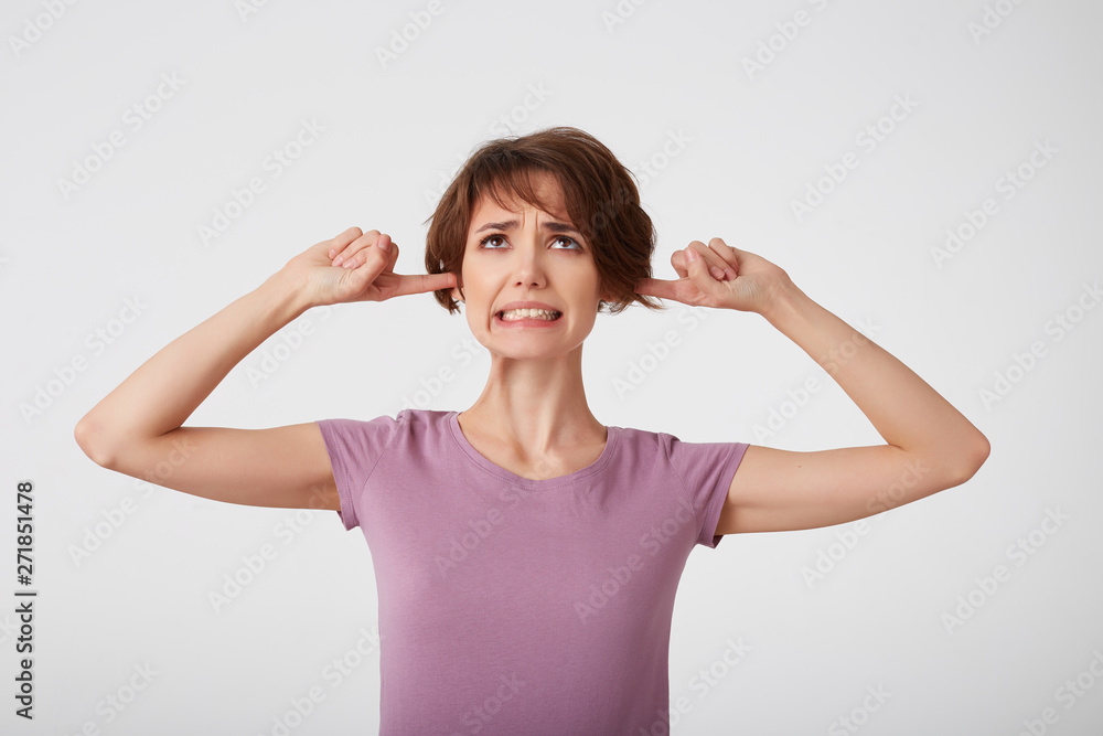 Young nice doubted short-haired lady in blank t-shirt, plugs ears as hears awful noise or music, squints face, avoids loud sound, ignores something unpleasant. Stands over white wall. Young nice doubted short-haired lady in blank t-shirt, plugs ears as hears awful noise or music, squints face, avoids loud sound, ignores something unpleasant. Stands over white wall.