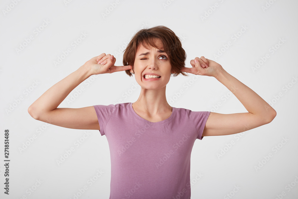 Fototapeta premium Young nice doubted short-haired lady in blank t-shirt, unwills hear bothering noise, plugs earholes with index fingers, clenches teeth and frowns face in dissatisfaction. Stands over white wall.
