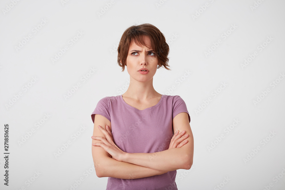 Portrait of frowning short-haired lady in blank t-shirt, trying to remember what offended her boyfriend, stands over white background with crossed arms. Portrait of frowning short-haired lady in blank t-shirt, trying to remember what offended her boyfriend, stands over white background with crossed arms.