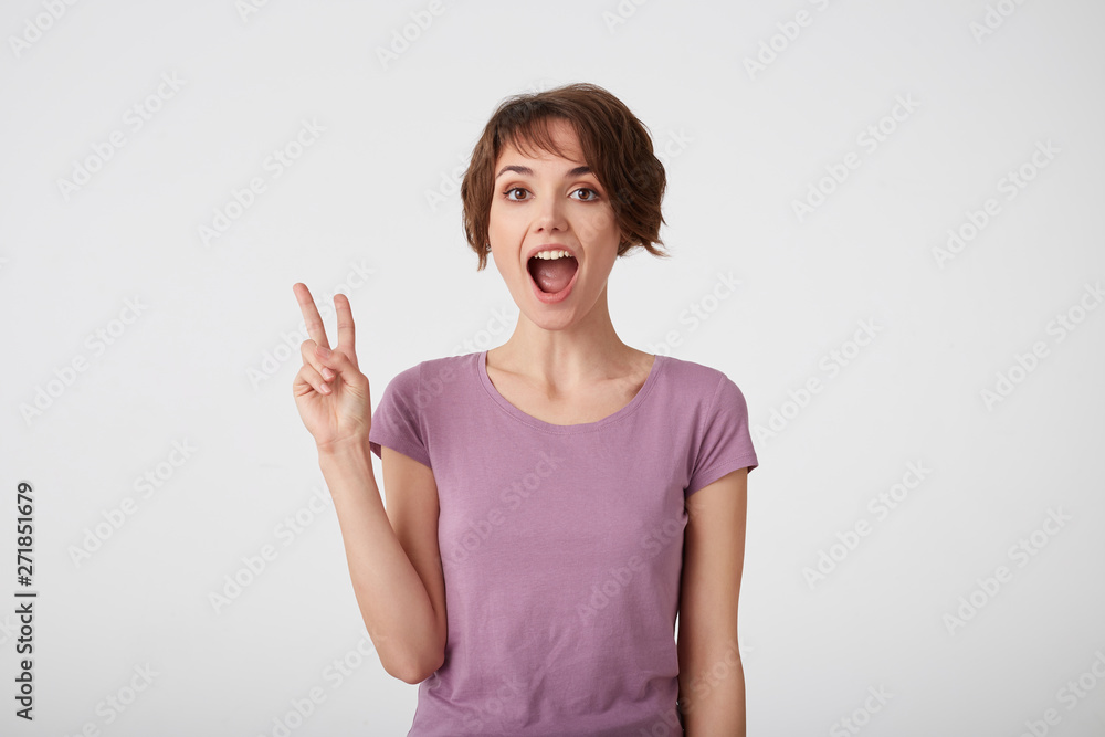 Funny young short-haired woman with wide open mout, makes peace sign with hand, expresses happiness, wears casual shirt poses indoor, stands over white wall.