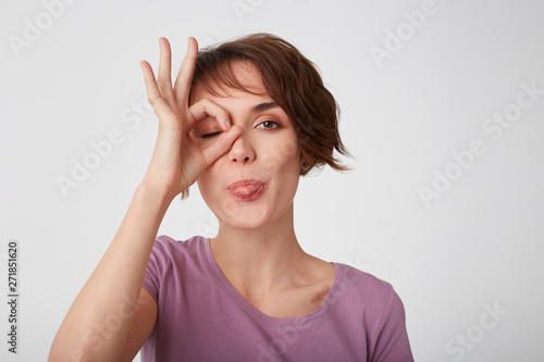 Wallpaper Mural Close up of young attractive funny short-haired lady in blank t-shirt, standing over white wall, looking through okay gesture and shows tongue. Positive emotion concept. Torontodigital.ca