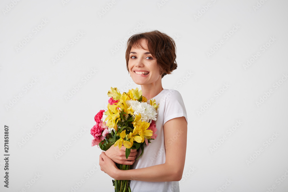 Young smiling cute short haired girl in white blank t-shirt, holding a bouquet of colorful flowers, very glad to such a gift from her boyfriend, bites his lip and smiling over white wall. Young smiling cute short haired girl in white blank t-shirt, holding a bouquet of colorful flowers, very glad to such a gift from her boyfriend, bites his lip and smiling over white wall.