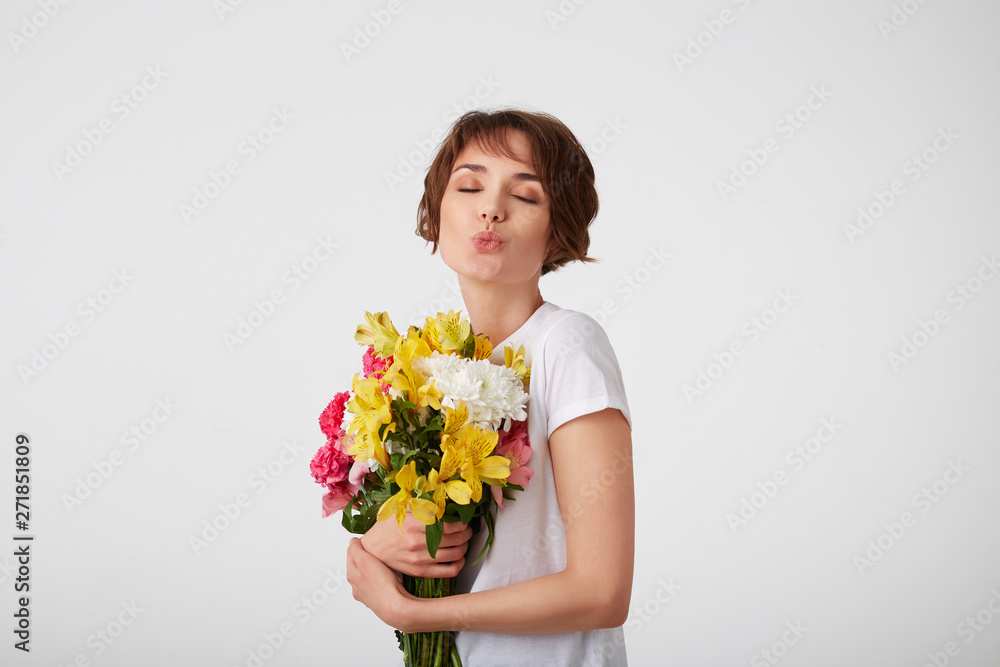 Portrait of young lovely cute short haired lady in white t-shirt, holding a bouquet of colorful flowers, very glad to such a gift from her boyfriend, sends a kiss with closed eyes over white wall. Portrait of young lovely cute short haired lady in white t-shirt, holding a bouquet of colorful flowers, very glad to such a gift from her boyfriend, sends a kiss with closed eyes over white wall.