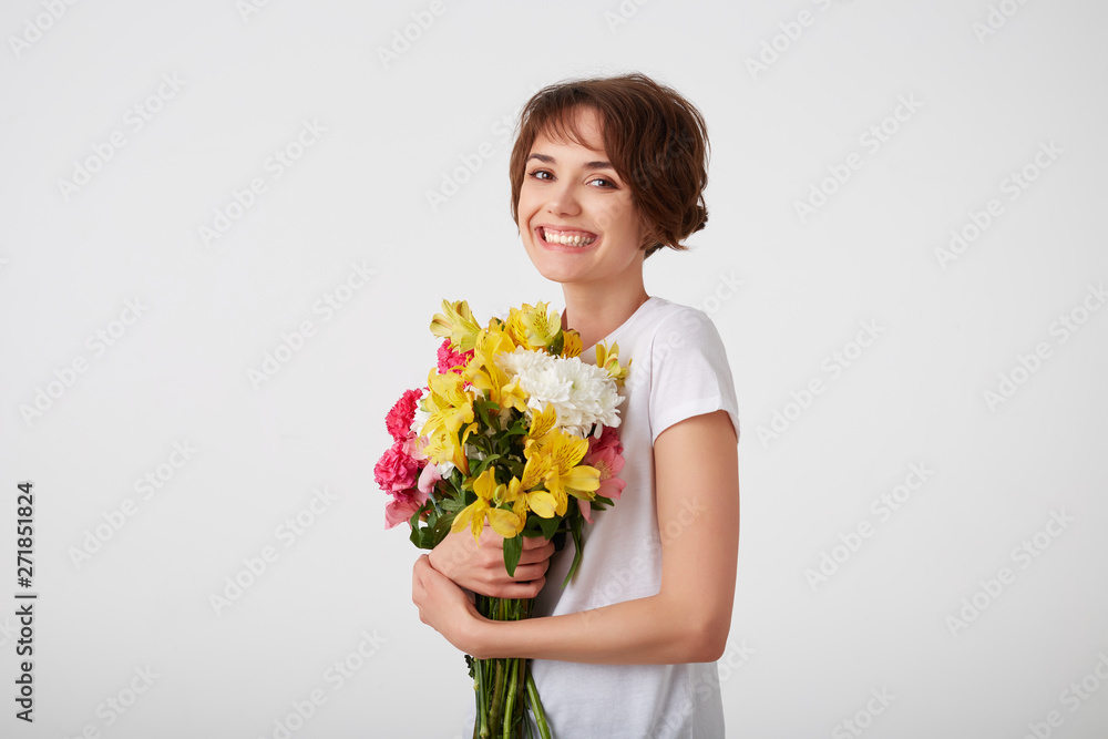 Portrait of young smiling cute short haired girl in white blank t-shirt, holding a bouquet of colorful flowers, very glad to such a gift from her boyfriend, broadly smiling over white wall. Portrait of young smiling cute short haired girl in white blank t-shirt, holding a bouquet of colorful flowers, very glad to such a gift from her boyfriend, broadly smiling over white wall.