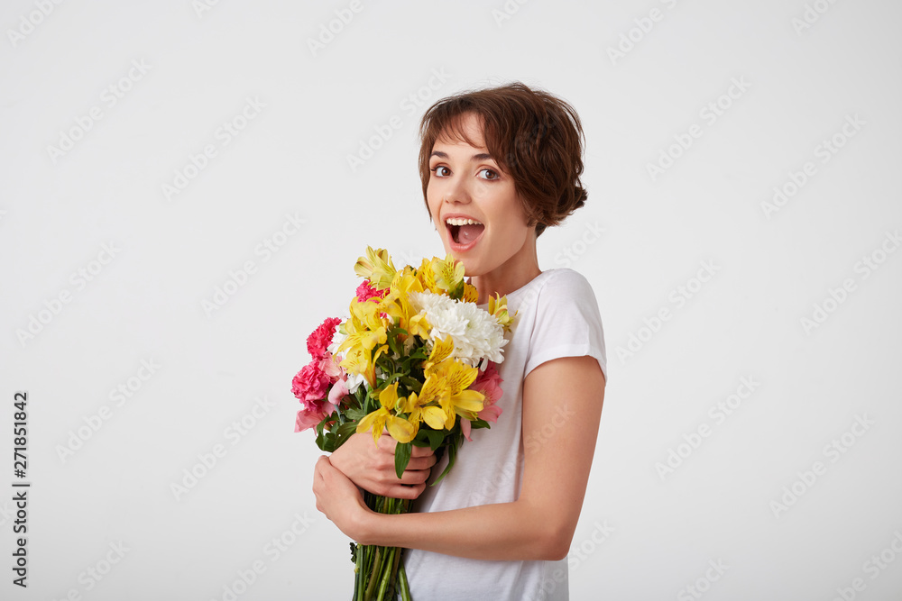 Young happy amazed cute short haired girl in white blank t-shirt, with wide open mouth and eyes, holding a bouquet of colorful flowers, surprised looking at the camera isolated over white wall.