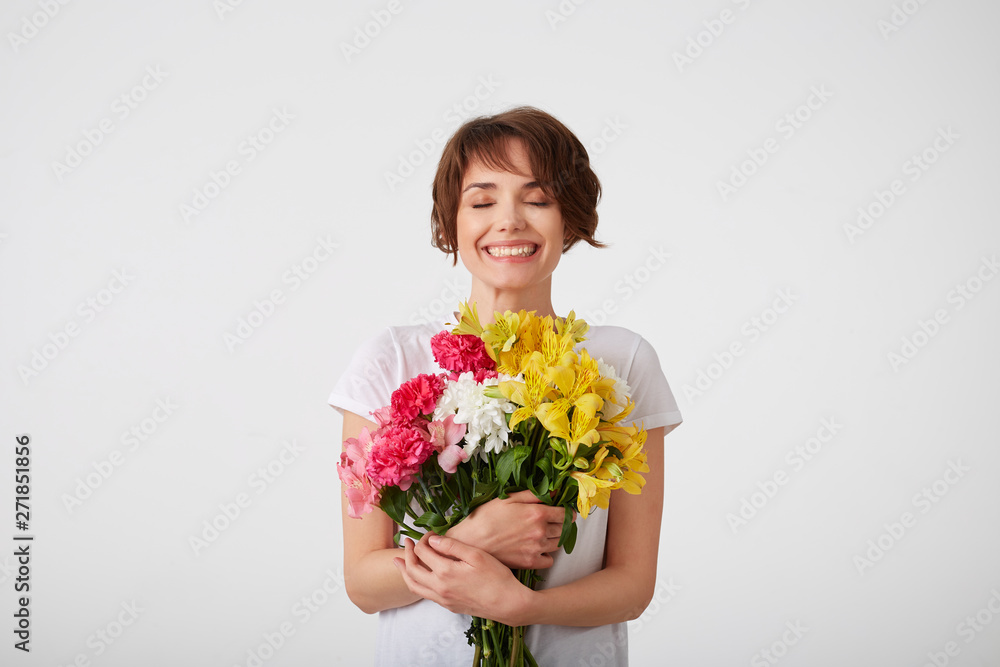 Happy cute young short haired girl in white blank t-shirt, holding a bouquet of colorful flowers with closed eyes, smiling broadly, standing over white background. Happy cute young short haired girl in white blank t-shirt, holding a bouquet of colorful flowers with closed eyes, smiling broadly, standing over white background.