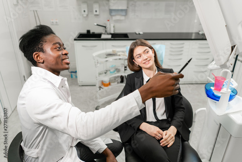 Young African male dentist shows x-rays on the monitor. Woman in the dentist chair at dental clinic. Medicine  health  stomatology concept. dentist treating a patient.
