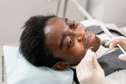 Young woman dentist treating root canals in the dental clinic. Young African American male with bad teeth lying on dentist chair with open mouth.