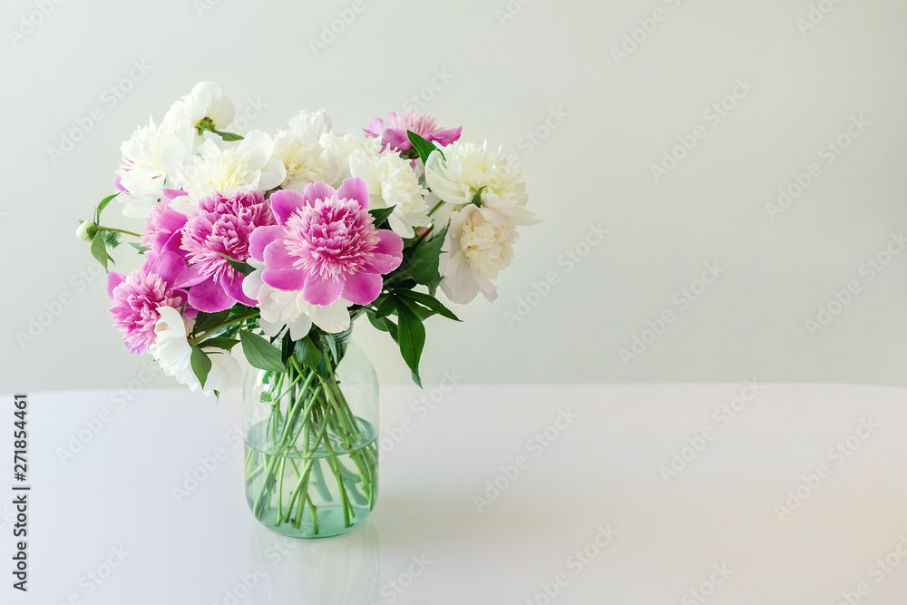 Bouquet of fresh big pink, white and cream peonies in simple glass jar on glance table indoor. Vase with beautiful tender spring flowers on glass table