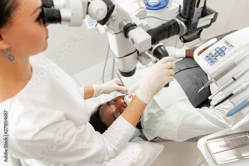 Modern equipment microscope in dental office. Young woman dentist treating root canals. Man patient lying on dentist chair with open mouth. Medicine, dentistry and health care concept.