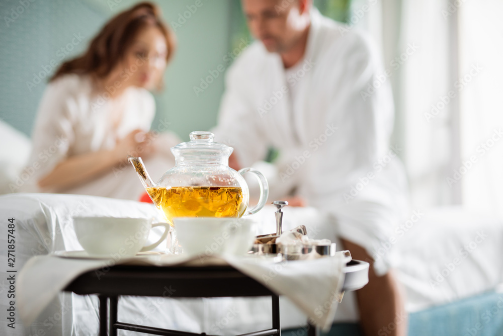 Glass teapot with two cups and couple sitting on the background Glass teapot with two cups and couple sitting on the background