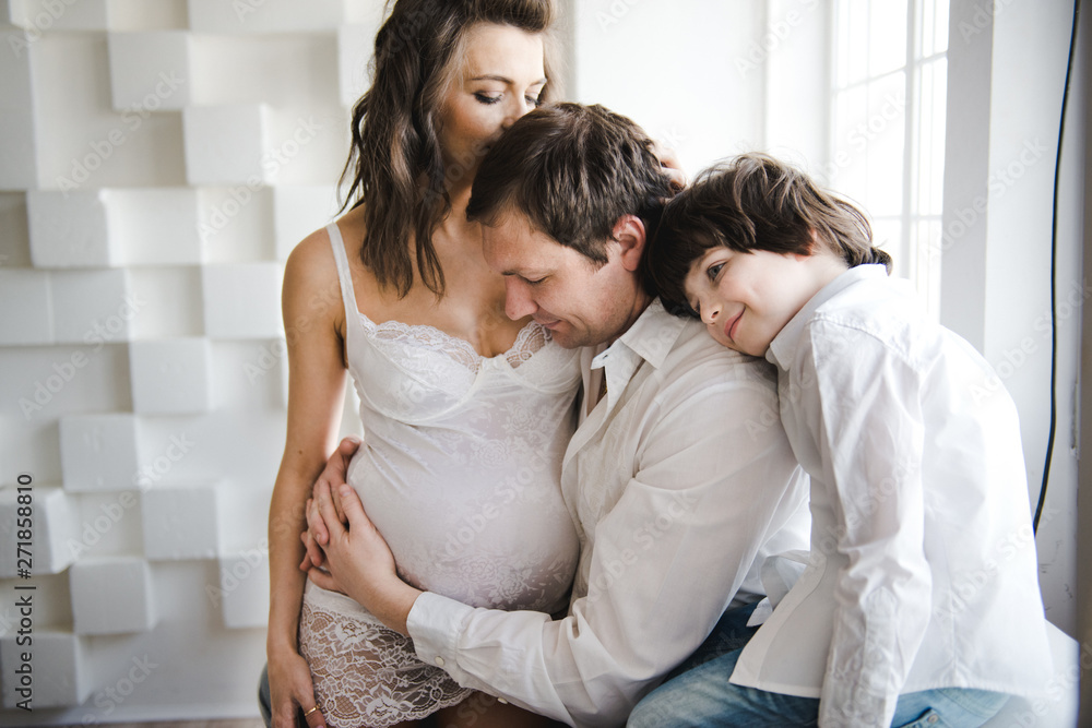 Happy family waiting for a second child. Pregnant woman with husband and little boy . Second pregnancy, maternity concept. Happy family near the window. White background. Happy family waiting for a second child. Pregnant woman with husband and little boy . Second pregnancy, maternity concept. Happy family near the window. White background.