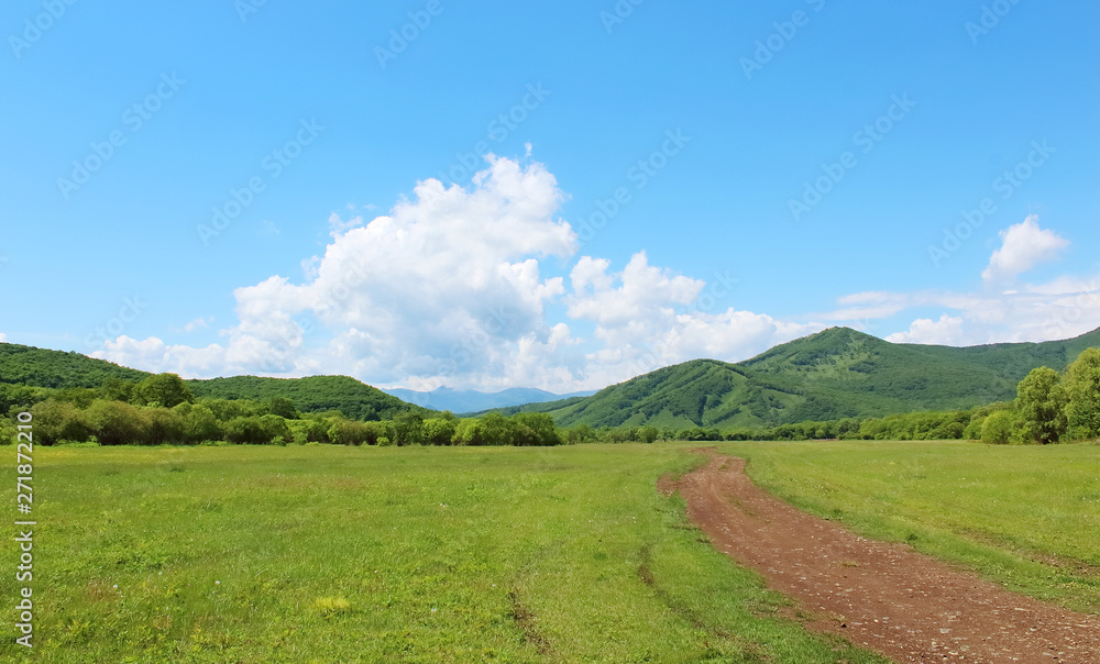 Summer landscape with green grass, hills, road and clouds