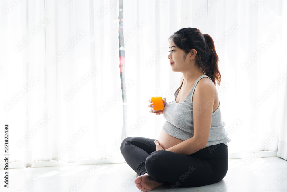 Asian pregnant woman holding a glass of orange juice and sitting near window, lifestyle concept. Asian pregnant woman holding a glass of orange juice and sitting near window, lifestyle concept.