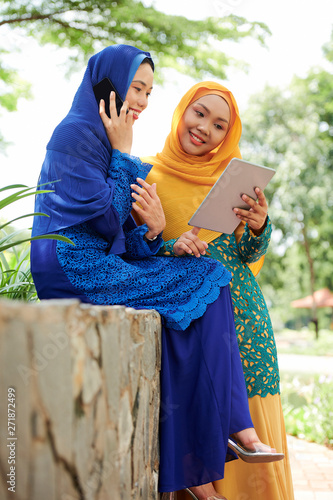 Smiling young woman in hijab shwing something on tablet computer to her friend talking on phone photo