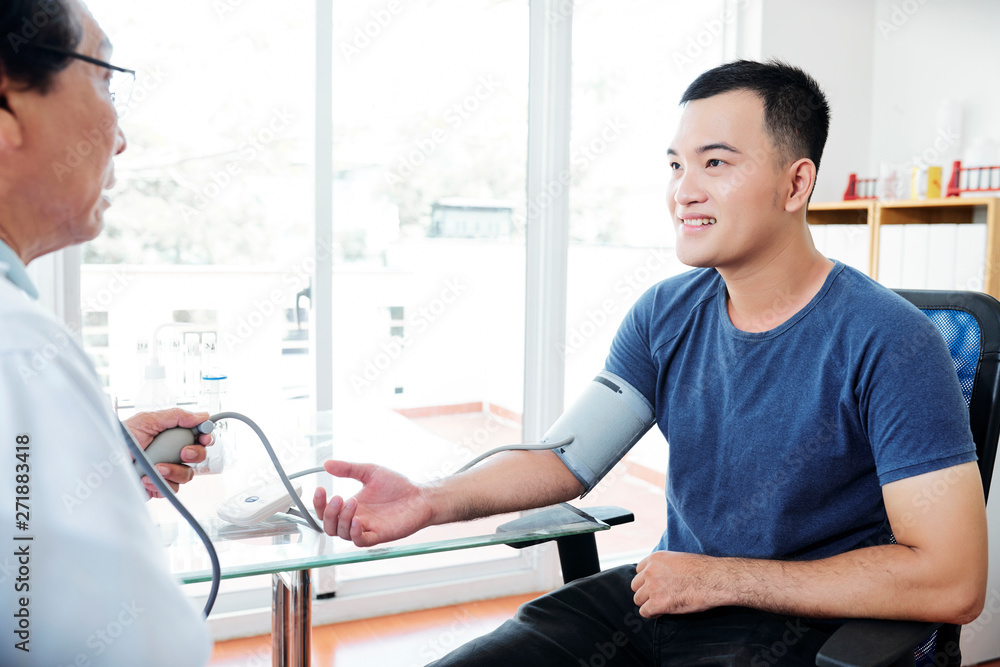 Fototapeta premium Doctor measuring blood pressure of young smiling Asian man