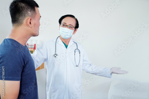 Senior general practitoner in medical mask asking patient to sit on bed