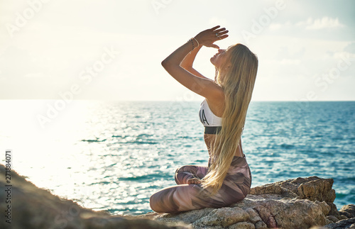 Young blonde woman doing yoga on the cliff by the sea at sunrise     