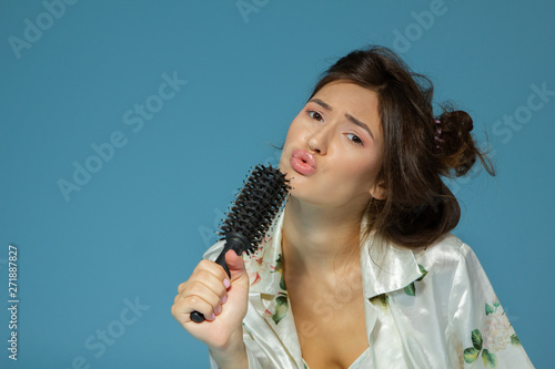 Cheerful attractive teen girl sing song holding comb like a microphone in the morning, over blue background.