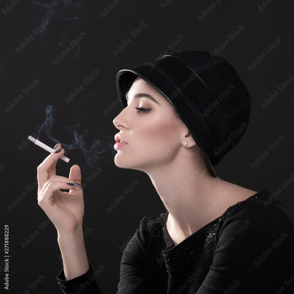 Obraz premium Young fashion woman smoking cigarette in hat and black drees over dark background. Vintage female portrait, styling. Image toned.