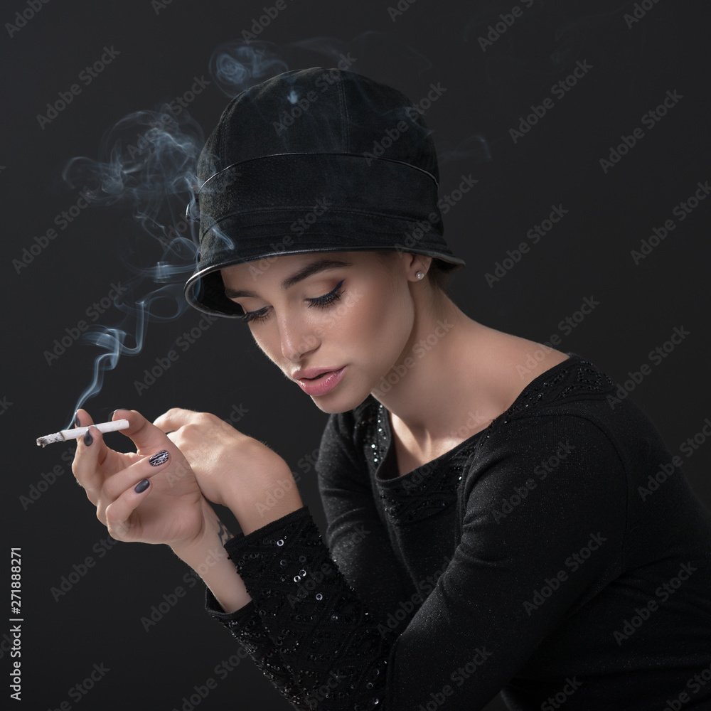 Obraz premium Young fashion woman smoking cigarette in hat and black drees over dark background. Vintage female portrait, styling. Image toned.