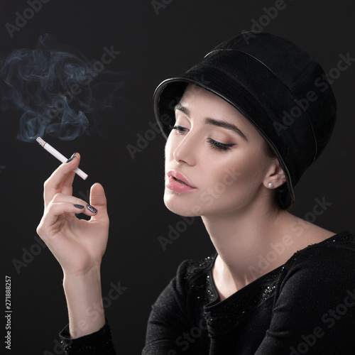 Young fashion woman smoking cigarette in hat and black drees over dark background. Vintage female portrait, styling. Image toned.