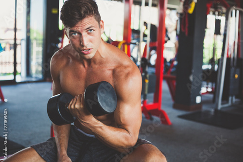handsome mole model with tanned muscular naked torso sits on a bench in the gym and trains biceps with dumbbells
