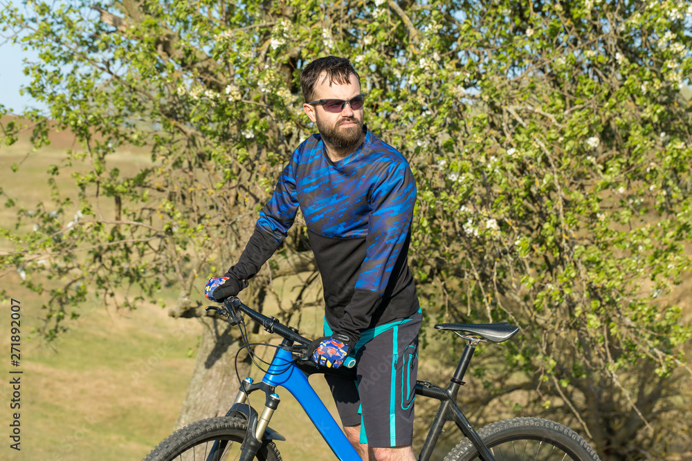 Cyclist in shorts and jersey on a modern carbon hardtail bike with an air suspension fork rides off-road on green hills near the forest	