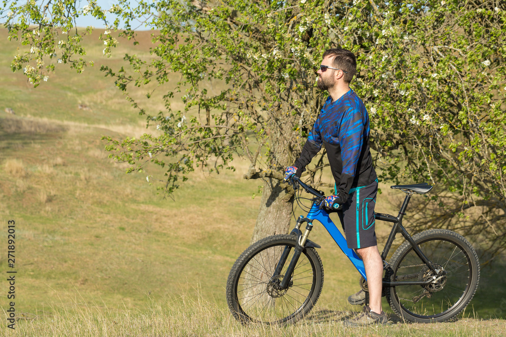 Cyclist in shorts and jersey on a modern carbon hardtail bike with an air suspension fork rides off-road on green hills near the forest	