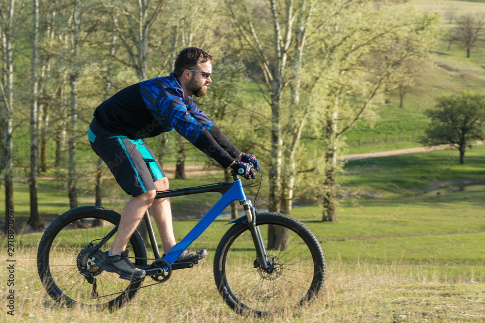 Cyclist in shorts and jersey on a modern carbon hardtail bike with an air suspension fork rides off-road on green hills near the forest Cyclist in shorts and jersey on a modern carbon hardtail bike with an air suspension fork rides off-road on green hills near the forest