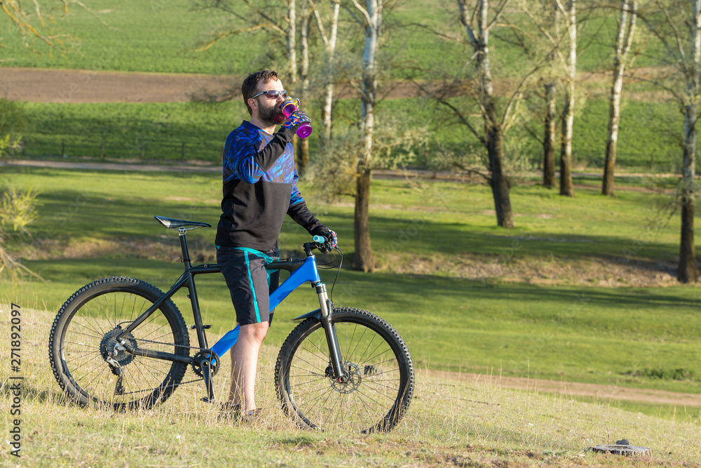 Cyclist in shorts and jersey on a modern carbon hardtail bike with an air suspension fork rides off-road on green hills near the forest	