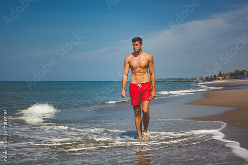 handsome sexy man in red shorts and with naked tanned fit torso walks on beach along the ocean. bright blue water and sky