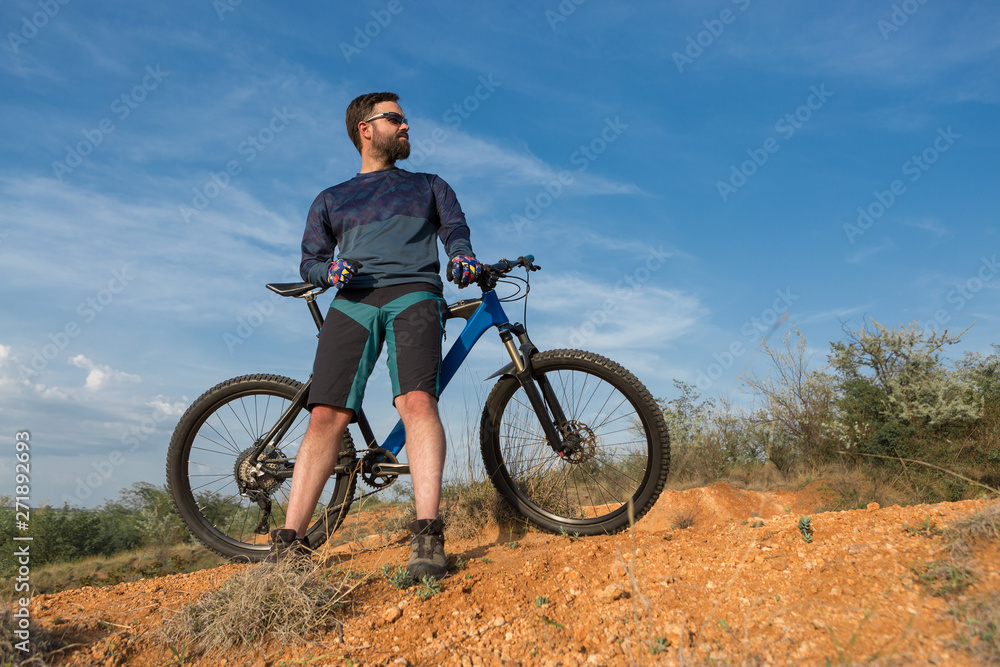 Cyclist in shorts and jersey on a modern carbon hardtail bike with an air suspension fork rides off-road on the orange-red hills at sunset evening in summer Cyclist in shorts and jersey on a modern carbon hardtail bike with an air suspension fork rides off-road on the orange-red hills at sunset evening in summer