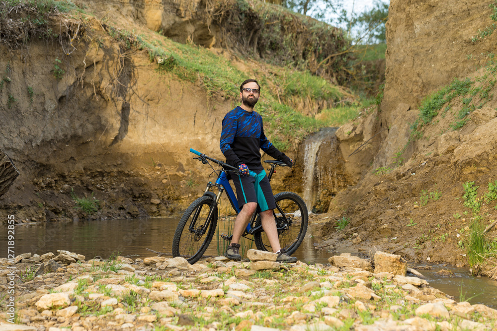 Cyclist in shorts and jersey on a modern carbon hardtail bike with an air suspension fork stands on the rocky shore of a mountain river Cyclist in shorts and jersey on a modern carbon hardtail bike with an air suspension fork stands on the rocky shore of a mountain river