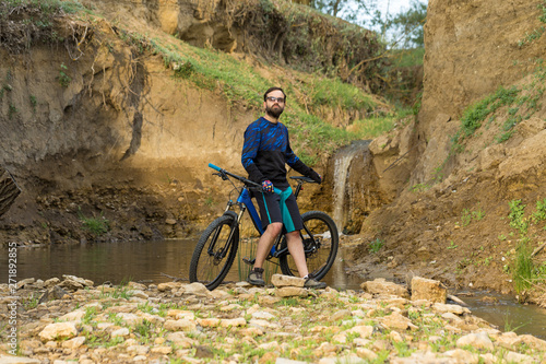 Cyclist in shorts and jersey on a modern carbon hardtail bike with an air suspension fork stands on the rocky shore of a mountain river  © Mountains Hunter