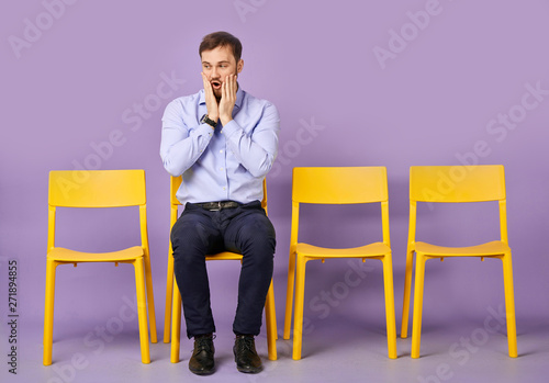 studio shot of astonished and amazed young man sitting on chair and looking aside and waiting for job interview