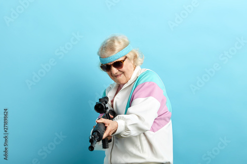 Senior woman in sports wear and black sanglasses screaming while aiming at camera with a rifle isolated on blue.
