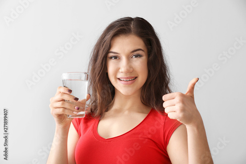 Beautiful young woman with glass of water showing thumb-up on light background