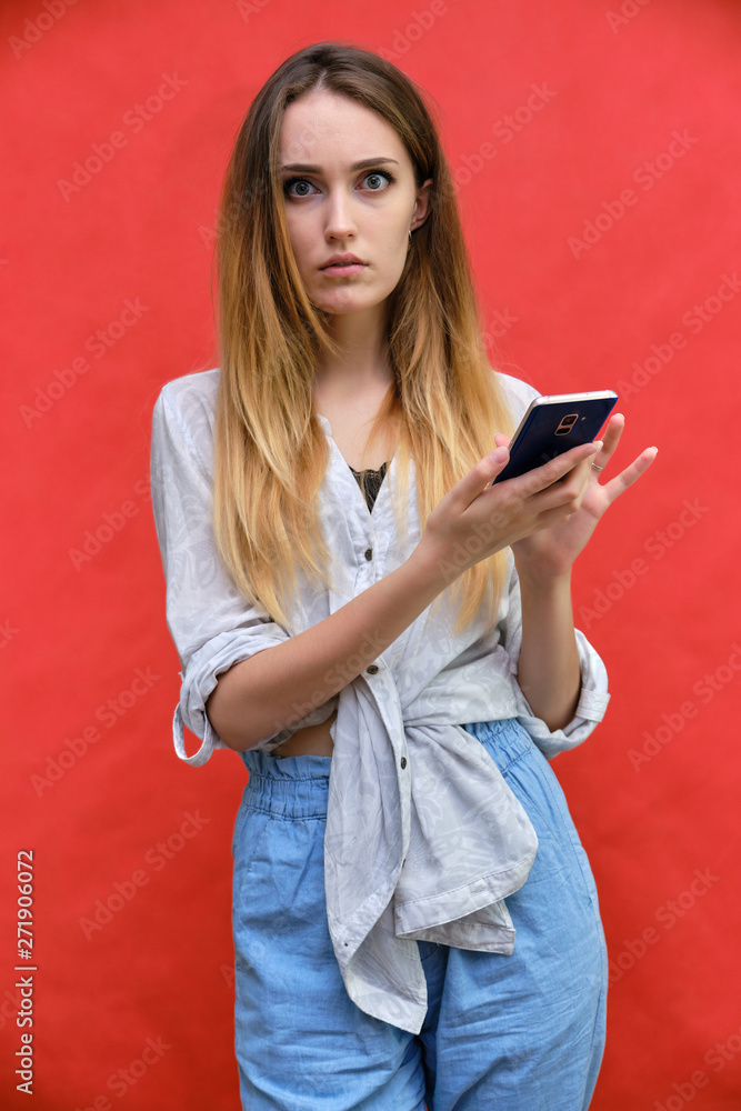 Naklejka premium Photograph of a portrait of a beautiful fashionable beauty girl woman with long dark flowing hair, on a red background with a smartphone. She is standing in different poses and smiling.
