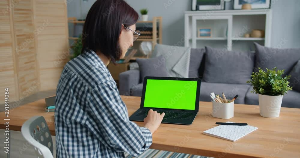 Smiling girl is using laptop with green screen working with computer at home sitting at table looking at mock-up monitor. People and communication concept. Smiling girl is using laptop with green screen working with computer at home sitting at table looking at mock-up monitor. People and communication concept.