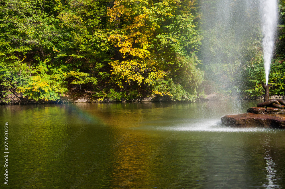 Water jet fountain on rock in center of lake with colorful fall trees reflected in calm water surface. Rainbow in water flow. Landscape of Sofiyivka park in Uman Ukraine. Autumn in Sofievka Uman park 